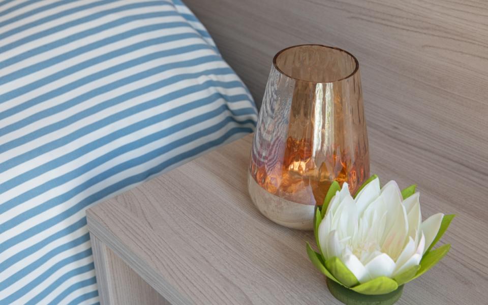 Comodino con vaso ambrato e fiore bianco, accanto a cuscino a righe blu.
