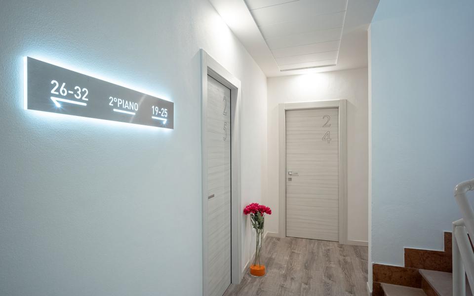 Corridoio moderno con porte numerate, segnaletica luminosa e fiori decorativi.