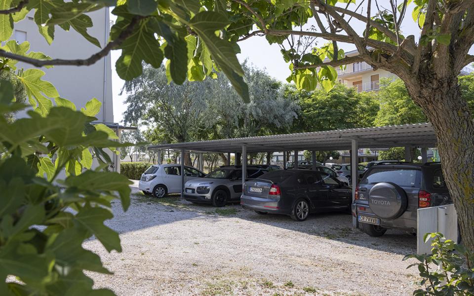 Parcheggio coperto con auto, alberi e palazzi sullo sfondo, giornata soleggiata.