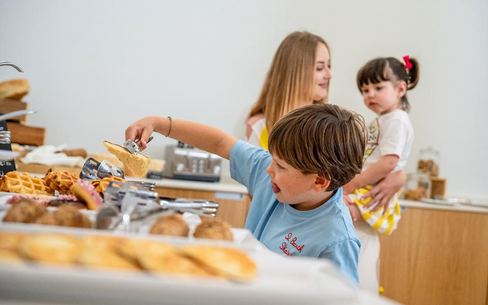 Bambino prende cibo da buffet, donna tiene una bambina in braccio.