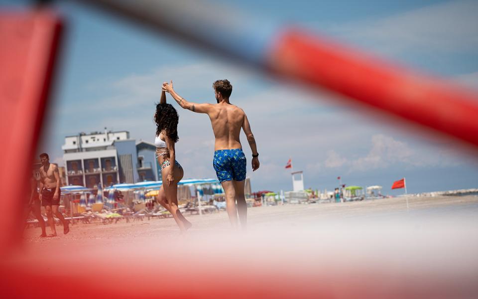 Coppia balla sulla spiaggia, ombrelloni e bandiere rosse sullo sfondo.